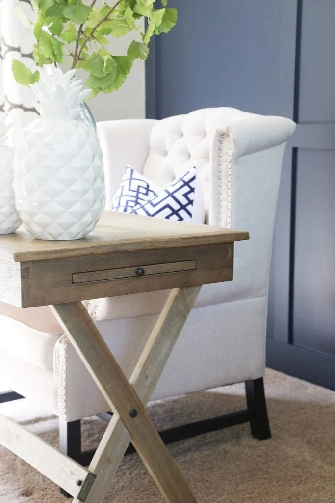 A white tufted armchair in front of the desk with a white vase on the desk and branches of green leaves in the vase.