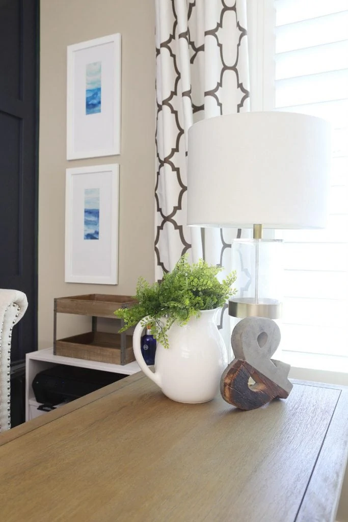 On the office desk is a white vase, a wooden & sign and a small white and glass table lamp.