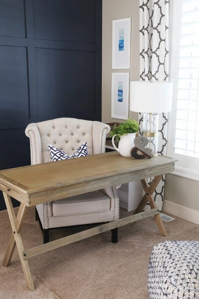 A wooden desk with an off white armchair in front of it and a navy wall behind it. These is a white pitcher with greenery on the desk.