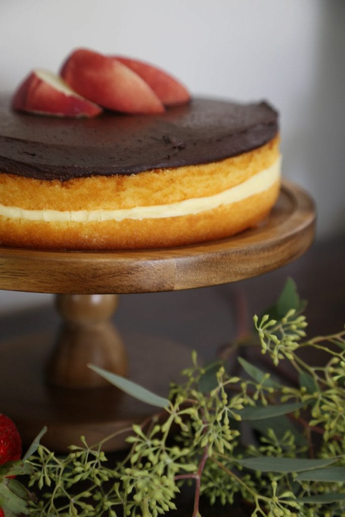 Wooden cake stand with sponge cake, chocolate frosting on it.