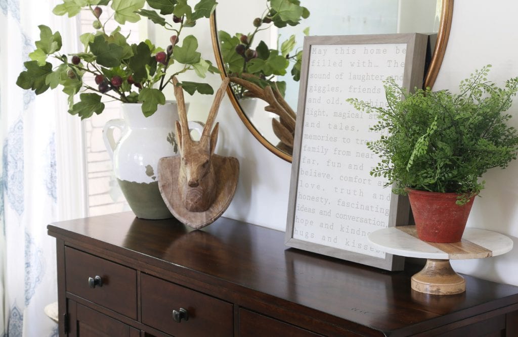 Dark wood buffet with a vase and green leaves in it, plus a plant on a cake stand.