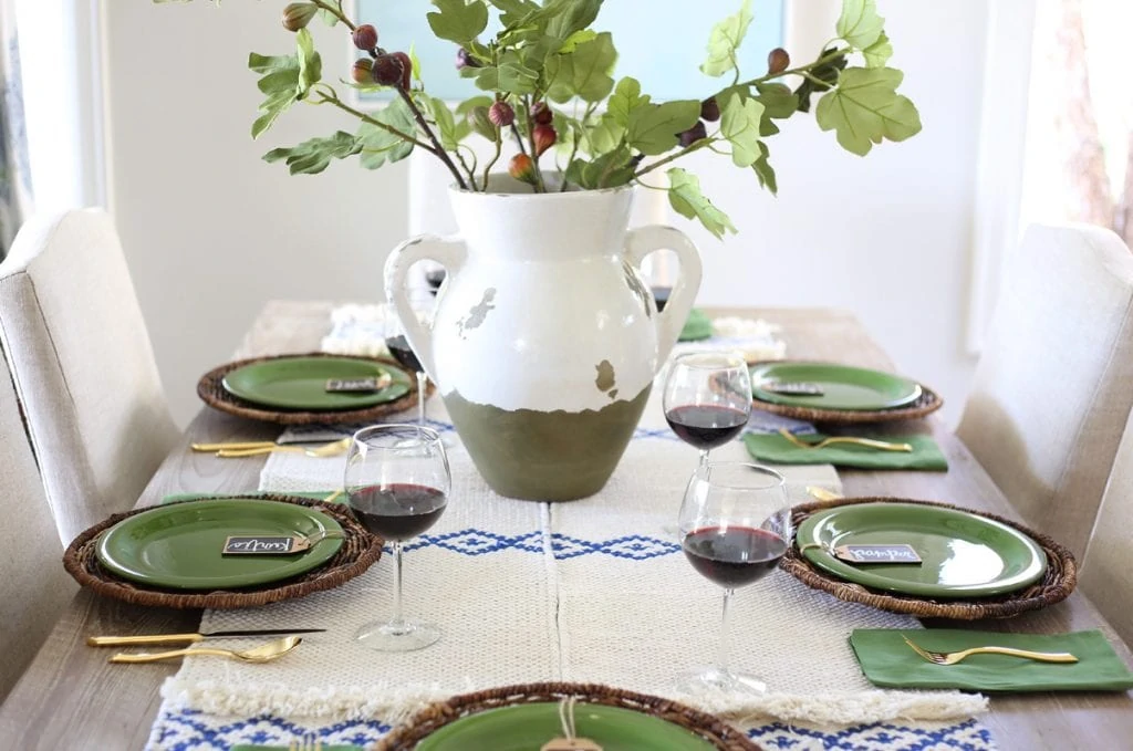 Dining room table with green plates and a blue and white runner. The centerpiece is a white and green vase with green leaves in it.