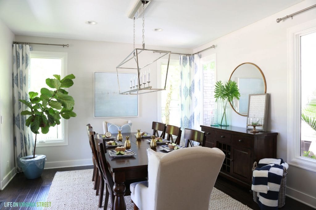 A dining room with a wooden table and chairs with the head table chairs in a white fabric.   There is a potted plant in the corner of the room and a large pendant light above the table.