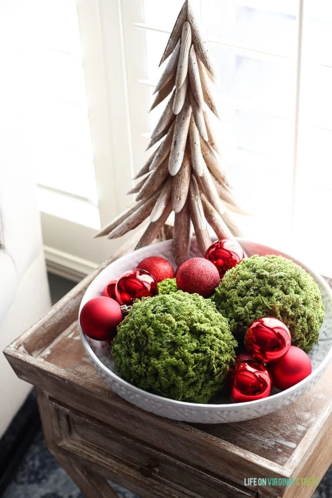 Decorative bowl filled with moss balls, and red ornaments, and a wooden tree beside it all on the side table.