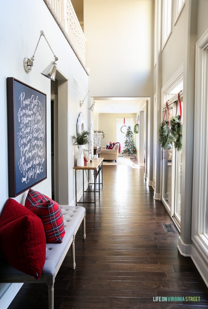 Christmas entryway hallway with linen bench, red plaid pillows, wreaths, swing-arm sconces and 'Doxology' canvas artwork. 