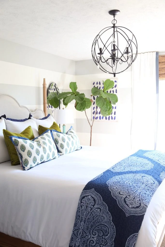 Bedroom with white bedding, striped walls, iron orb chandelier, and blue and green accents. Love the added greenery with the fiddle leaf fig tree.