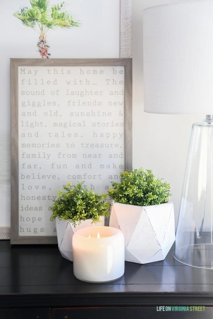 A lit candle on a wooden table with plants around it and a graphic word framed picture behind it.