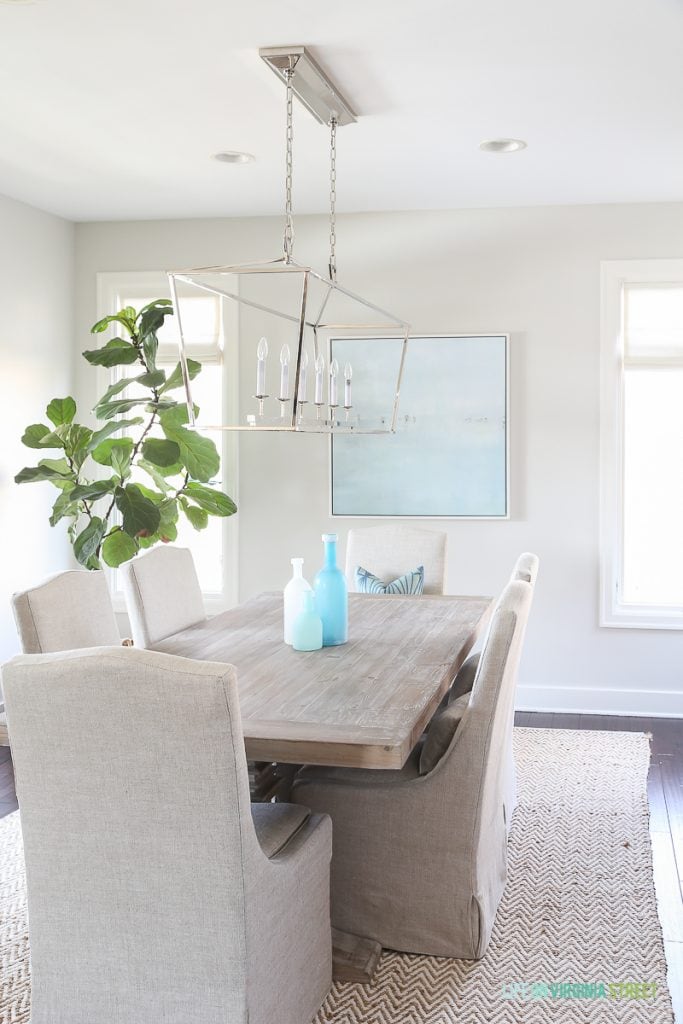 Coastal dining room with natural wood table, linen chairs, fiddle leaf fig tree, Darlana linear pendant and blue ocean art. Love the simple linen roman shades!