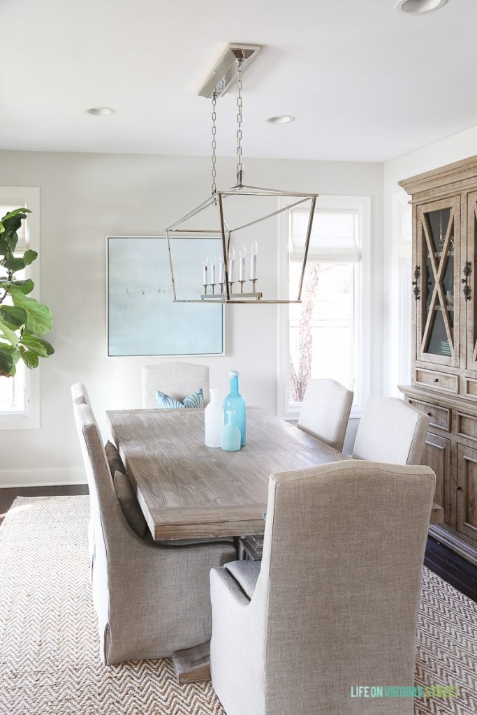 Coastal dining room with natural wood table, linen chairs, fiddle leaf fig tree, Darlana linear pendant and blue ocean art. Love our new dining room Roman shades!