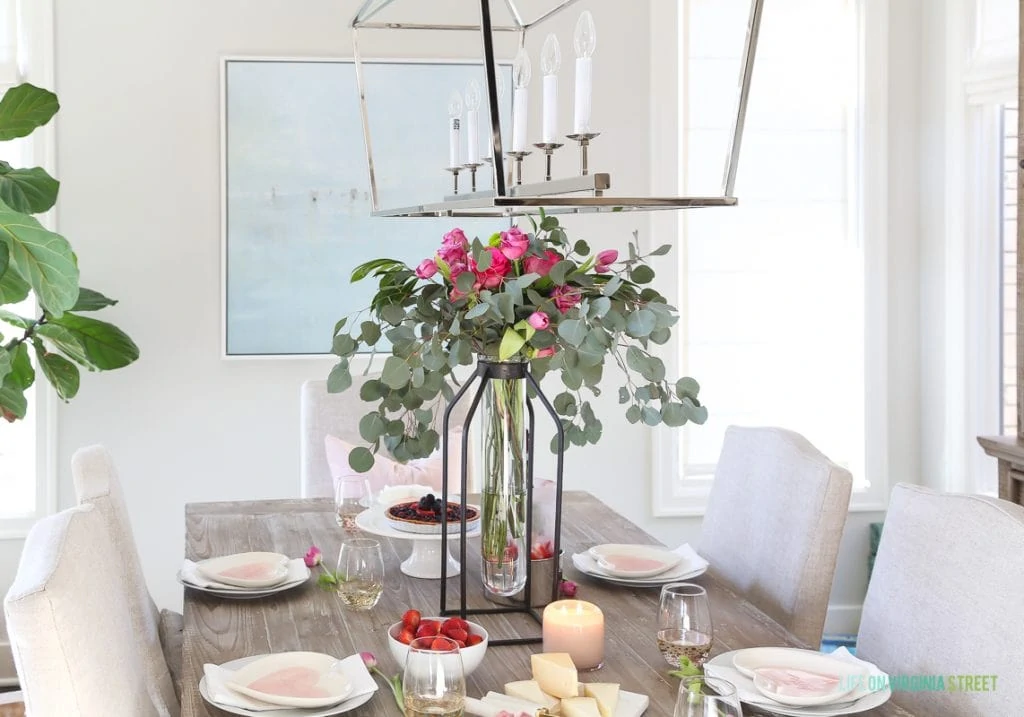 A tall floral and eucalyptus vase, linen chairs, reclaimed wood table, beach art, and heart plates on the dining room table.