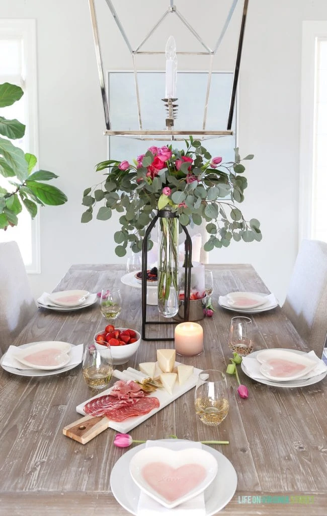 Valentine's Day entertaining and tablescape with tall floral and eucalyptus vase, linen chairs, reclaimed wood table, beach art, and heart plates. A fresh and colorful tablescape!