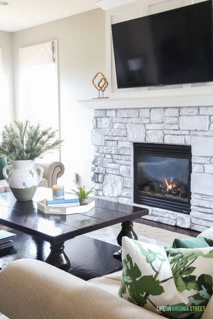  A neutral living room with a white washed fireplace that is on, a tv above the mantel, a wooden coffee table and leaf motif pillows on the couch.