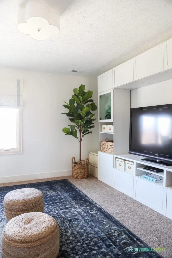 TV and craft room with Benjamin Moore Simply White walls, fiddle leaf fig tree, scalloped light fixture, blue vintage-style rug and sisal poufs.