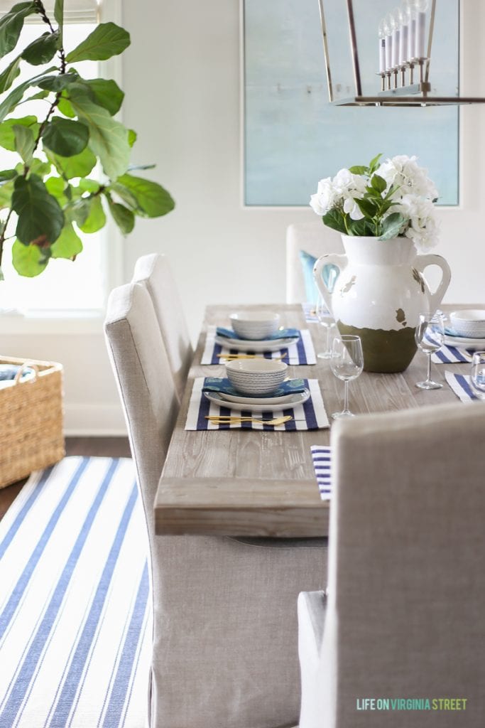 A white and blue striped rug underneath the dining room table, with a large potted green plant in the corner of the room.