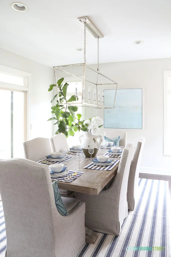 A coastal inspired tablescape with navy blue and white striped placemats, blue paisley napkins, blue squiggle dishes, white hydrangeas, beach art, linen dining chairs, reclaimed wood hutch, driftwood table, Darlana, linear pendant, fiddle leaf fig tree, gold silverware and blue striped rug.