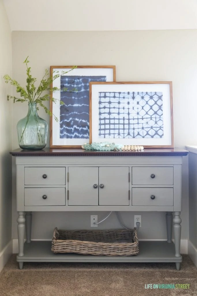 Gray buffet table dresser with indigo shibori artwork, glass demijohn, wood and glass beads, faux greenery, and woven basket. Walls are Behr Castle Path.