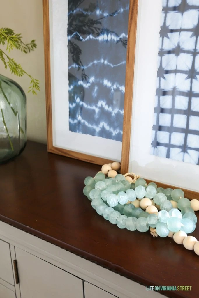 Gray buffet table dresser with indigo shibori artwork, glass demijohn, wood and glass beads, faux greenery, and woven basket. Walls are Behr Castle Path.