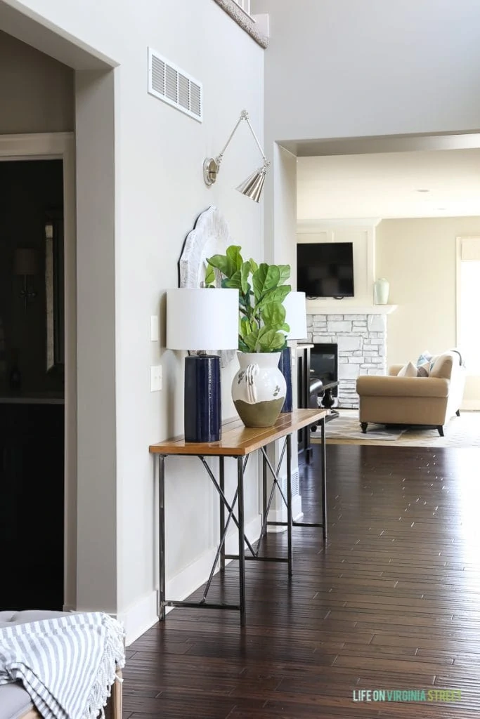 Gorgeous neutral entryway hallway with gray linen driftwood bench, gray and white striped throw, wood and iron console table, navy blue linen lamps, chrome swing-arm sconces, and faux fiddle leaf fig stems.