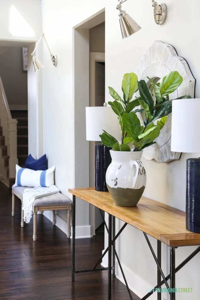 Gorgeous neutral entryway hallway with gray linen driftwood bench, gray and white striped throw, wood and iron console table, navy blue linen lamps, chrome swing-arm sconces, and faux fiddle leaf fig stems.