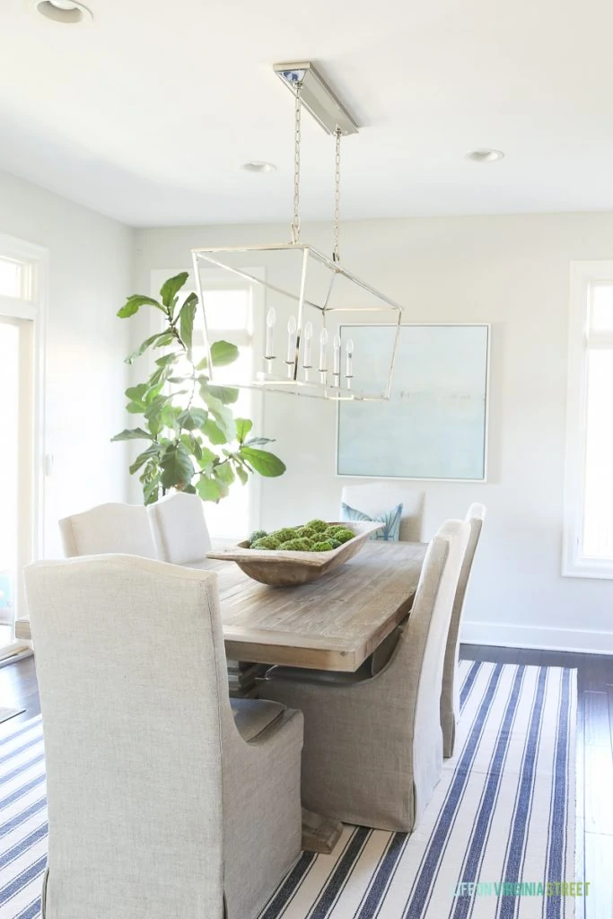 Coastal dining room with navy blue striped rug, beach artwork, fiddle leaf fig tree, dough bowl and reclaimed wood dining table.