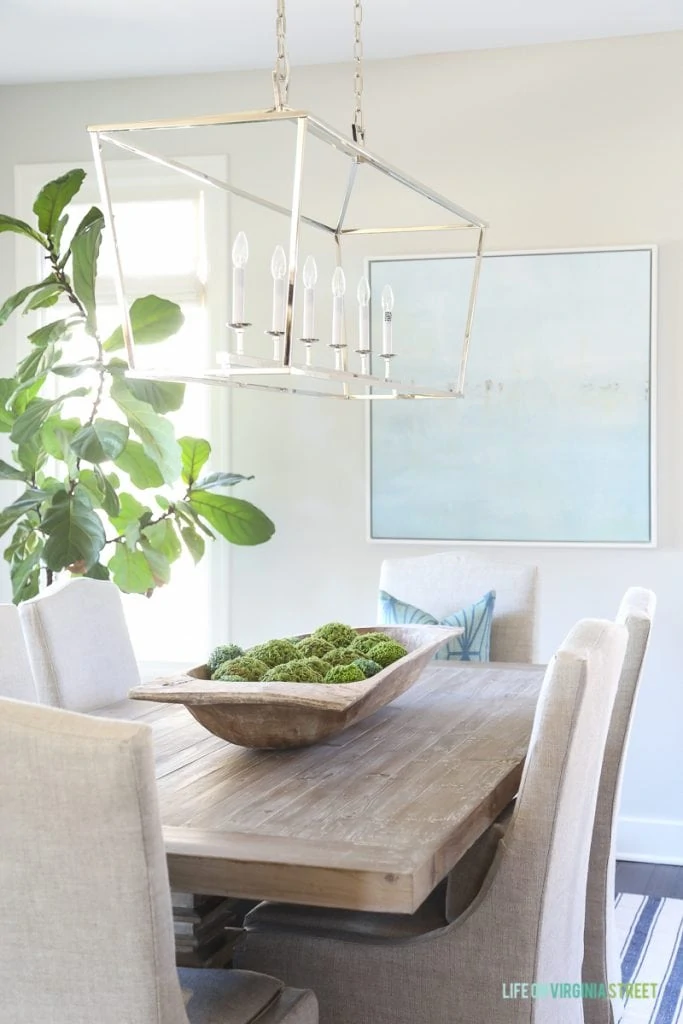 Coastal dining room with navy blue striped rug, beach artwork, fiddle leaf fig tree, dough bowl and reclaimed wood dining table.