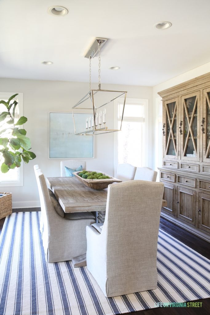 Coastal style dining room with navy blue and white striped rug, reclaimed wood hutch, driftwood color table, linen chairs, Darlana linear pendant light, fiddle leaf fig tree and beach art.