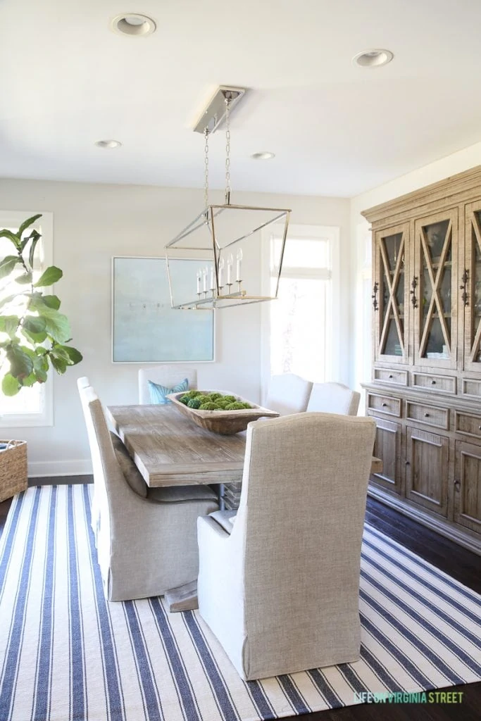 Coastal dining room with navy blue striped rug, beach artwork, fiddle leaf fig tree, dough bowl and reclaimed wood dining table.