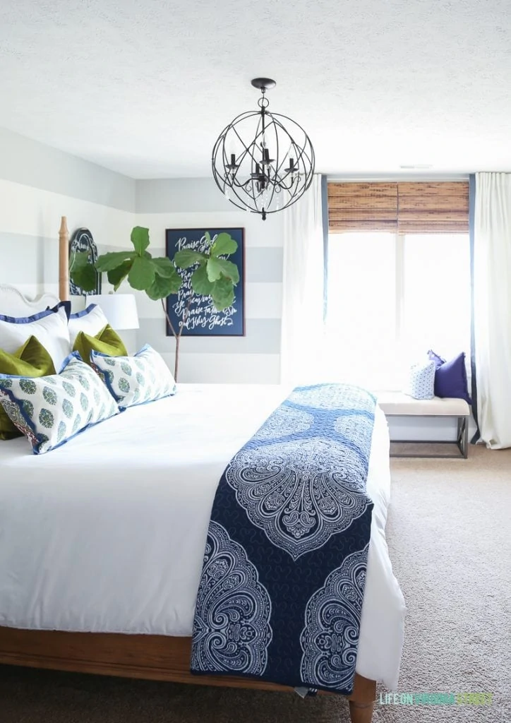 Guest bedroom with woven shades, ribbon trimmed drapes, white bedding, navy blue paisley throw, Doxology canvas, fiddle leaf fig tree, chartreuse velvet pillows and hickory wood bed.