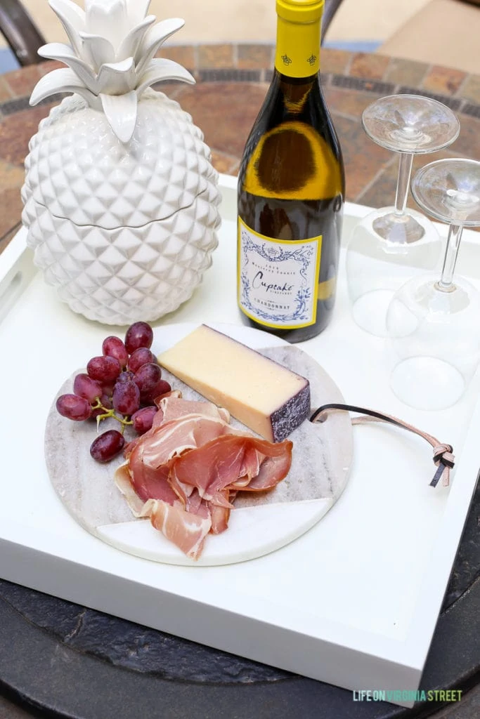 Marble cheese board with prosciutto, grapes, and cheese on a white serving tray. Love the white ceramic pineapple and the oleander tree in the background.