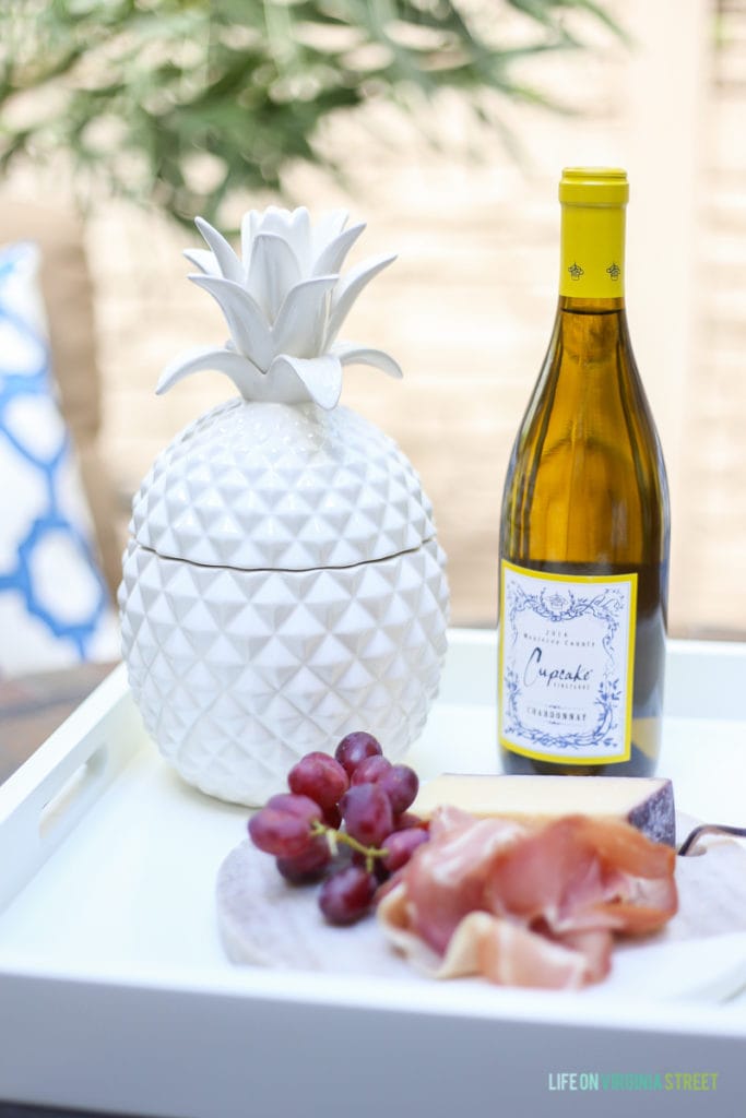 Marble cheese board with prosciutto, grapes, and cheese on a white serving tray. Love the white ceramic pineapple and the oleander tree in the background.