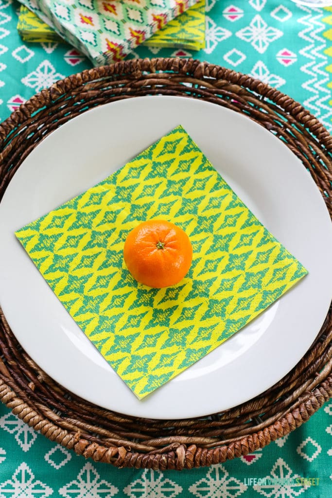 A white plate with a blue and yellow napkin and an orange on the napkin to hold it down in the wind.