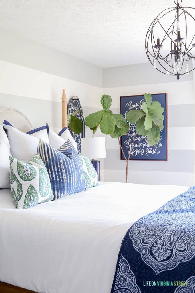 Bedroom with gray and white striped walls, white bedding, blue and green paisley pillows, navy blue linen lamps, iron orb chandelier and a navy blue Lindsay Letters Doxology canvas.