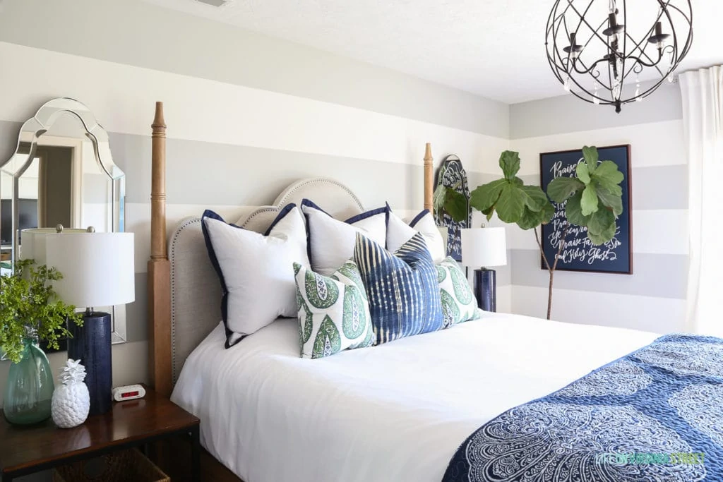 Side view featuring bedroom with gray and white striped walls, white bedding, blue and green paisley pillows, navy blue linen lamps, iron orb chandelier and a navy blue Lindsay Letters Doxology canvas.