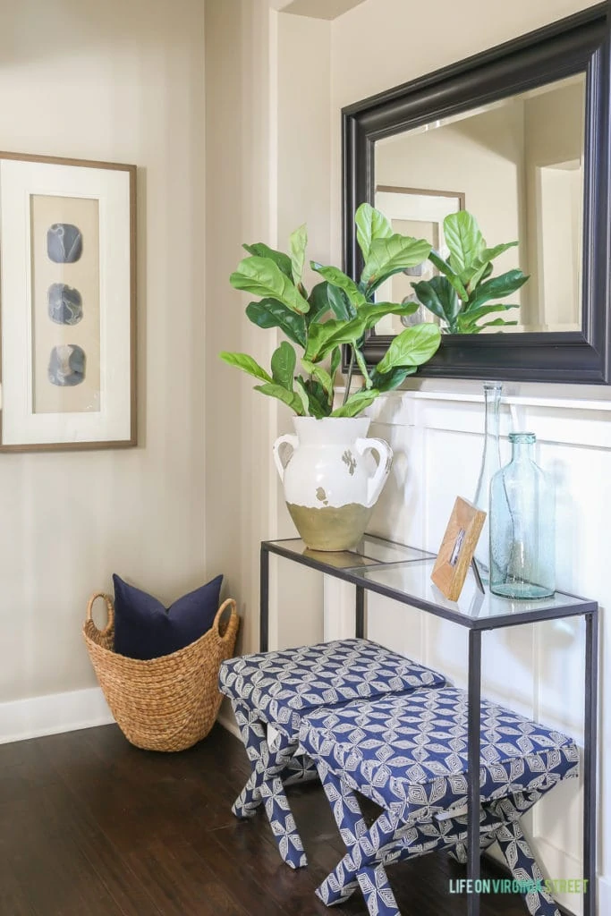 A beachy entryway vignette with blue and white patterned x-leg benches, seagrass basket, fiddle leaf fig stems, recycled glass vases, framed agate artwork, a wood frame, black mirror and navy blue velvet pillow.