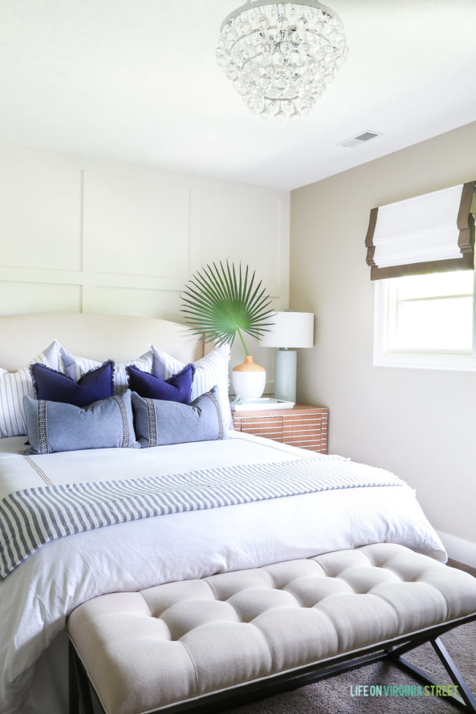 A coastal bedroom with white bedding, blue and white throw pillows, a linen bench, crystal semi flushmount chandelier, palmetto branch, white and wood vase, aqua lamp and ribbon-trimmed roman shades. Love that striped wood nightstand!