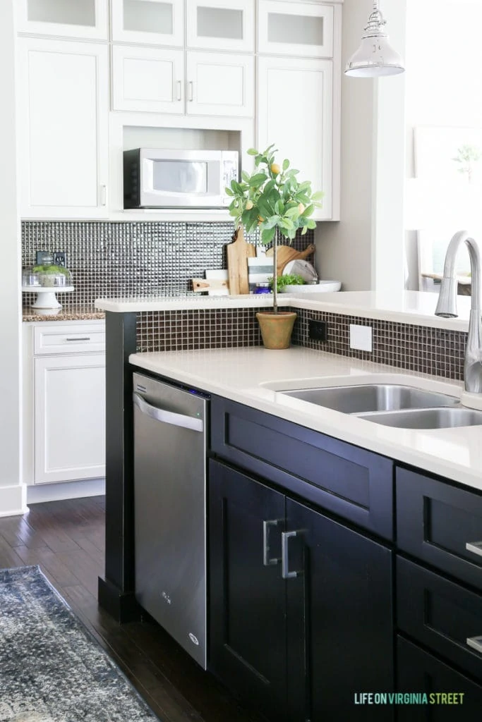A white kitchen with black island, quartz countertops, chrome pendants, and a blue and brown vintage-inspired rug.