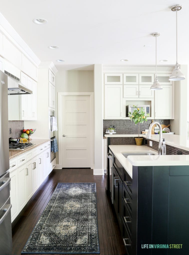 A white kitchen with black island, quartz countertops, chrome pendants, and a blue and brown vintage-inspired rug.