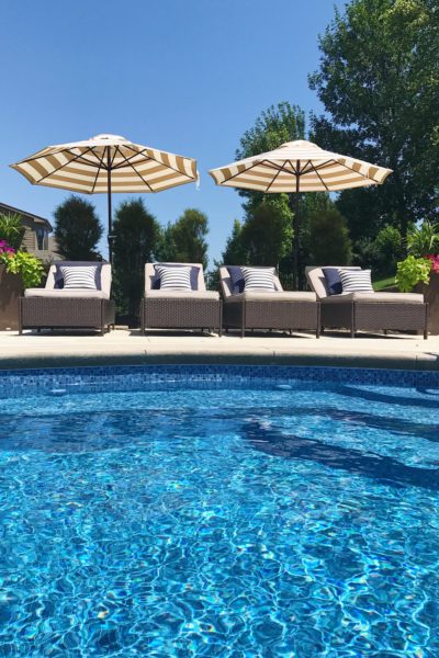 Blue vinyl pool with navy blue striped pillows on lounge chairs and beige and white striped umbrellas.