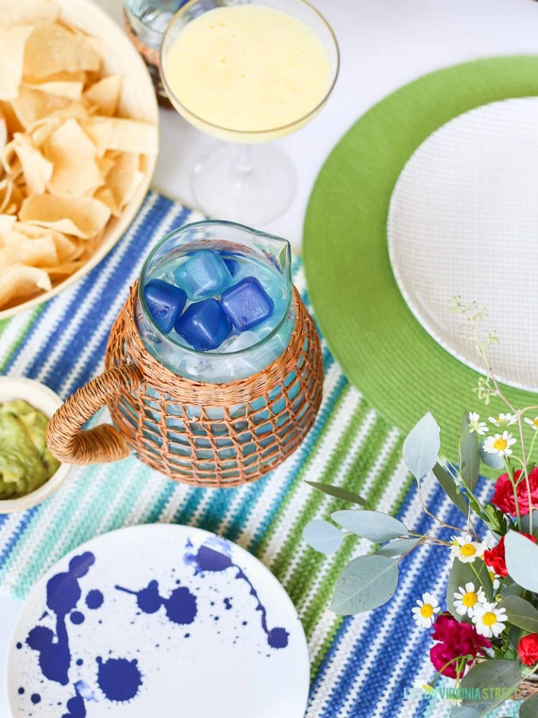 Rattan and glass water pitcher with reusable blue and aqua ice cubes. Blue, green and turquoise striped runner with splatter melamine plates.