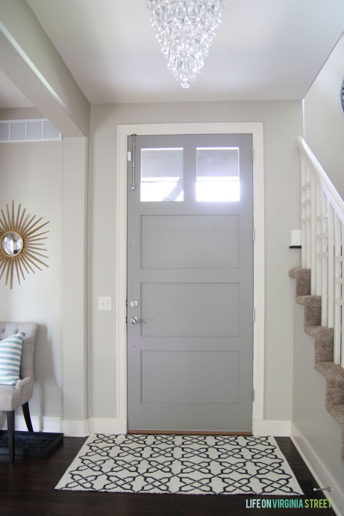 Entryway with gold starburst mirror, crystal teardrop chandelier, linen chair, navy blue trellis rug and blue and white striped pillow. Walls are Behr Castle Path and door is Behr Elephant Skin