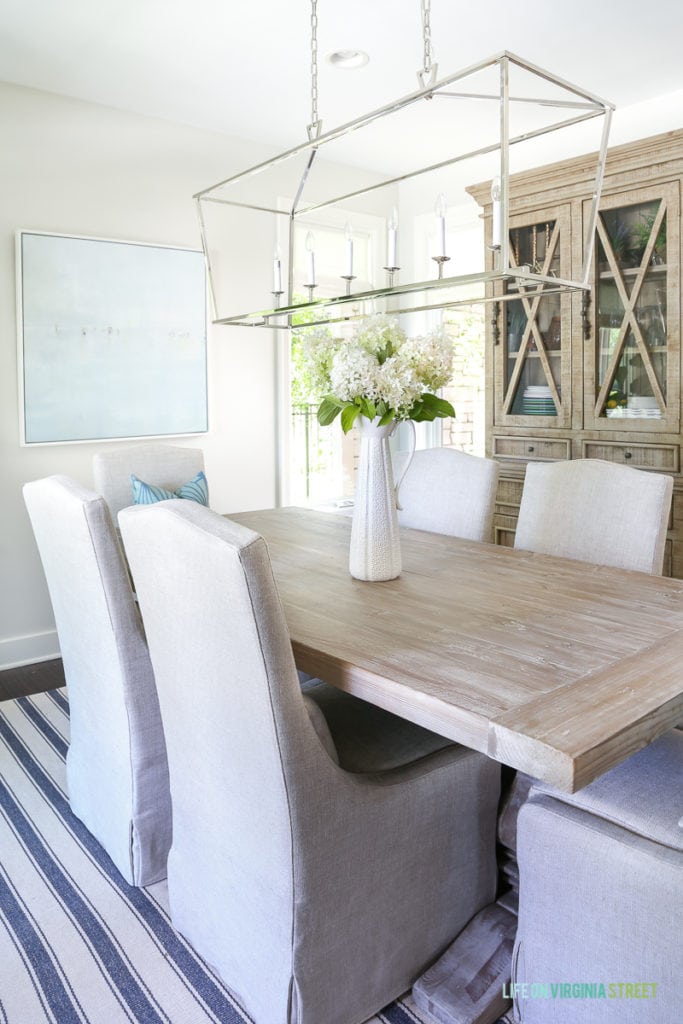 Light and bright dining room with blue and white striped rug, reclaimed wood dining table, linen slipcover dining chairs, Darlana linear pendant, beachy artwork, and fresh limelight hydrangeas. Wall color is Behr Silver Drop.