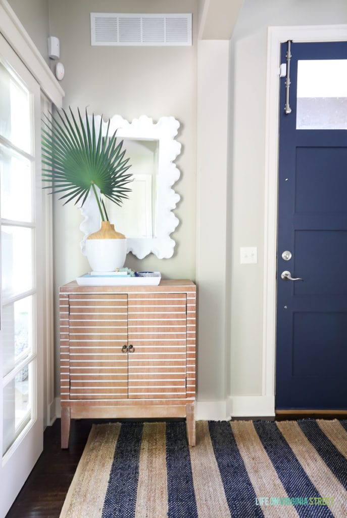 Coastal style entryway update with navy blue jute striped rug, striped wood cabinet, palm fronds, coral style mirror and Benjamin Moore Hale Navy blue painted interior door.