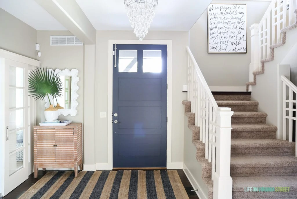 Coastal style entryway update with crystal teardrop chandelier, navy blue jute striped rug, striped wood cabinet, palm fronds, It Is Well canvas artwork from Lindsay Letters, coral style mirror and Benjamin Moore Hale Navy blue painted door.