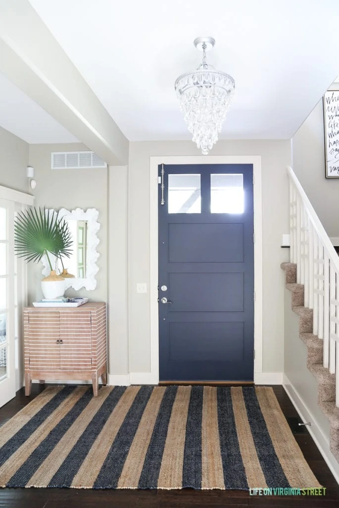 Coastal style entryway update with a painted interior door. Styled with a crystal teardrop chandelier, navy blue jute striped rug, striped wood cabinet, palm fronds, coral style mirror, Behr Castle Path walls, and Benjamin Moore Hale Navy blue painted door.