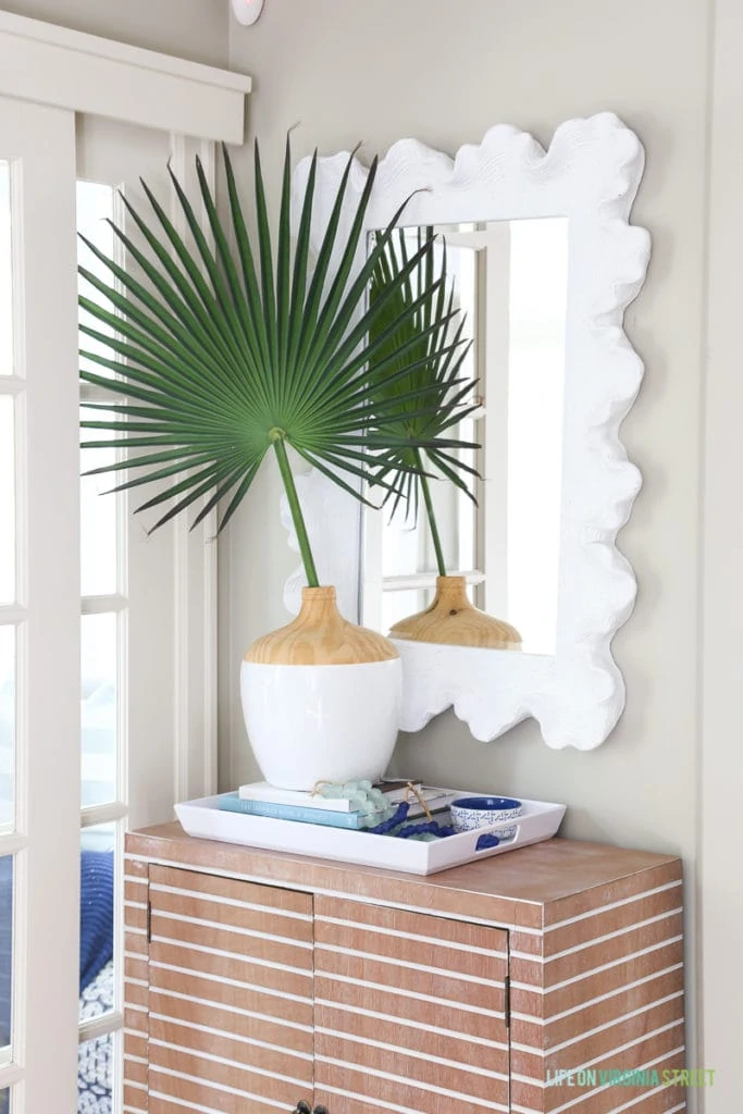 Coastal style entryway update with striped wood cabinet, tray with books and glass beads, palm fronds, coral style mirror.