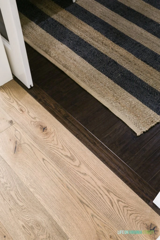 Light oak floors and dark wooden floors with a transition piece in between and striped rug on the floor.