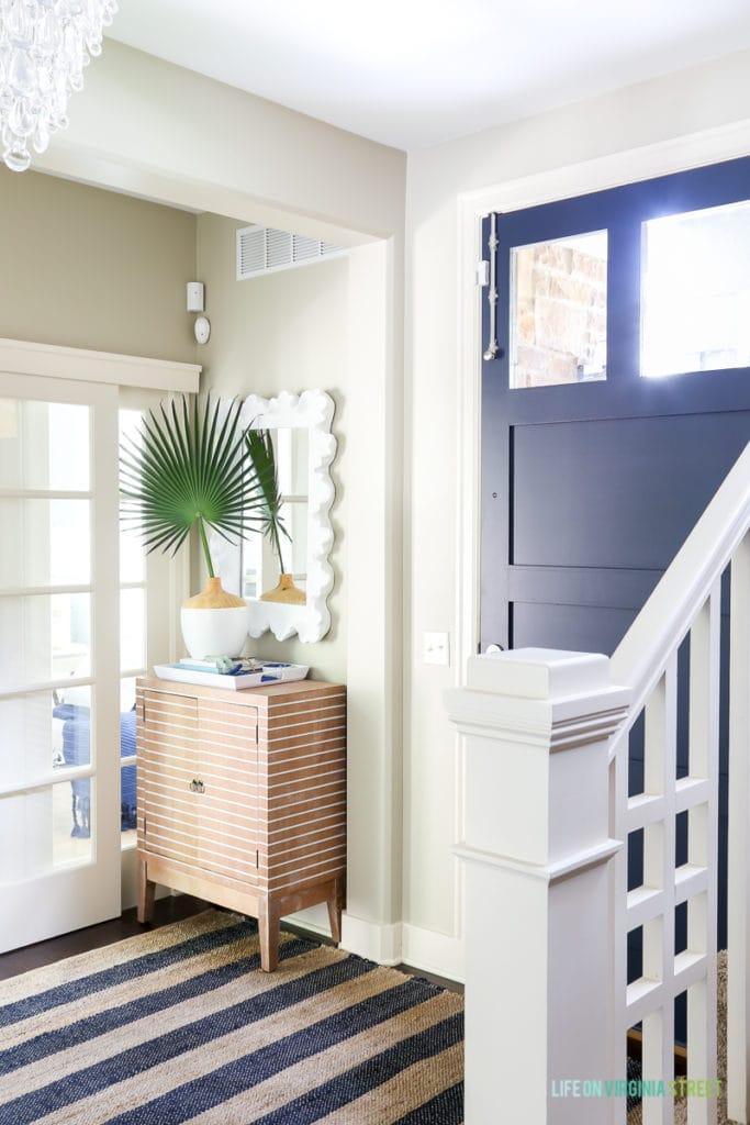 Coastal style entryway update with crystal teardrop chandelier, navy blue jute striped rug, striped wood cabinet, palm fronds, sliding french doors, coral style mirror and Benjamin Moore Hale Navy blue painted door.