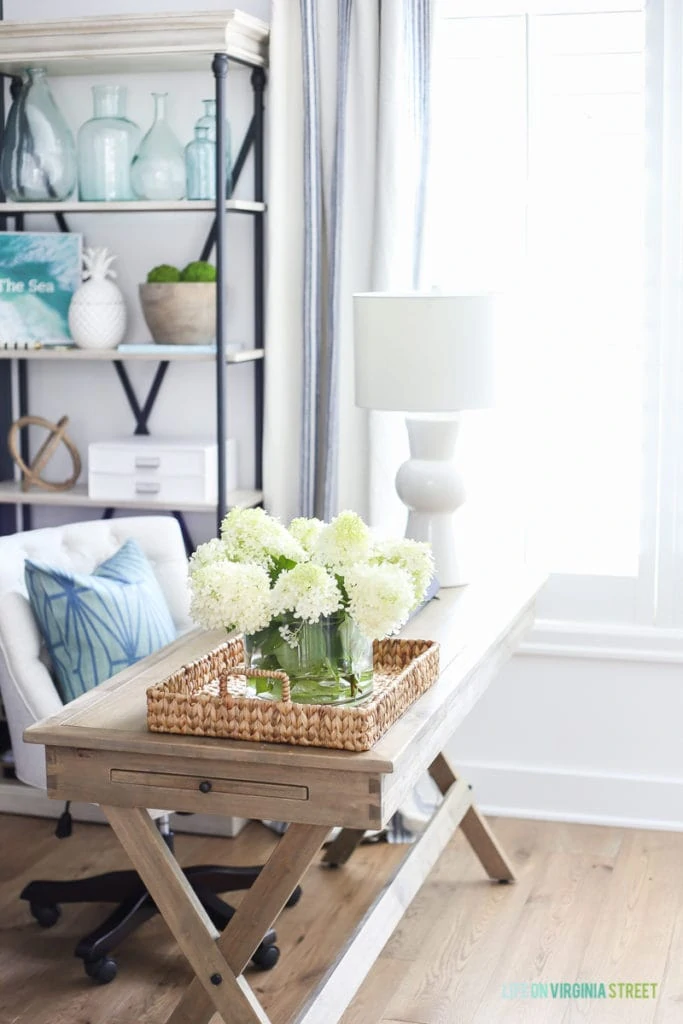 A wooden desk with a white office chair and a blue pillow. There are white flowers on the desk.