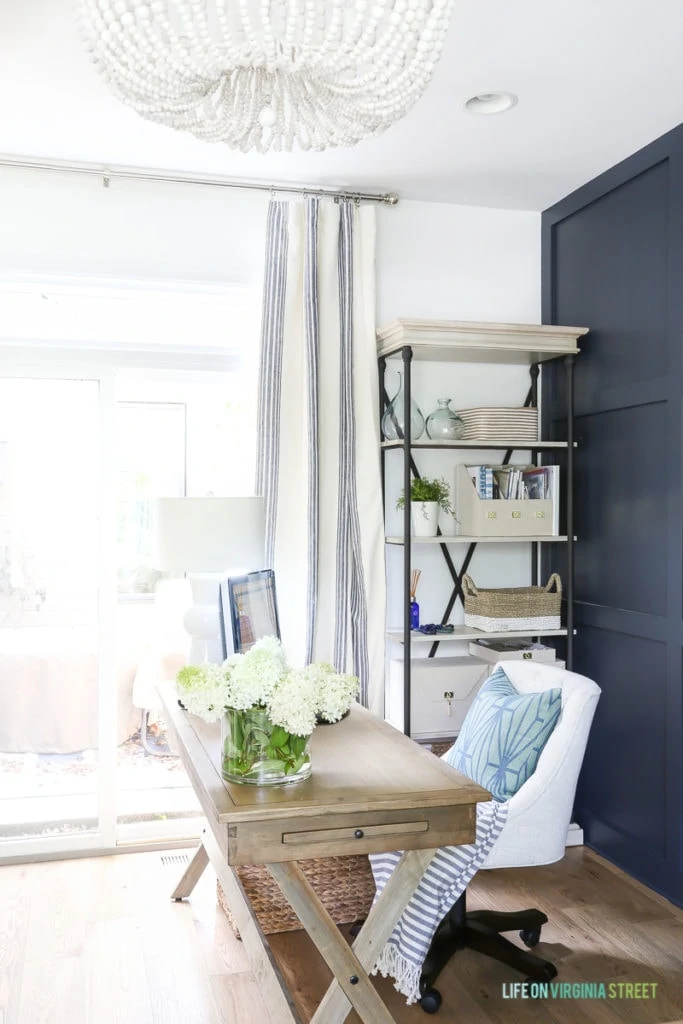 A wooden desk and chair in room and beside it a open metal shelf with office items on it.