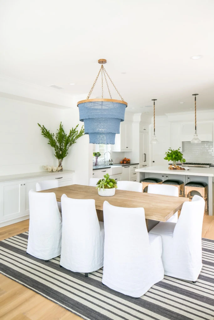 White dining room with natural wood table, white linen chairs, black and white striped rug, blue chandelier and fresh greenery.